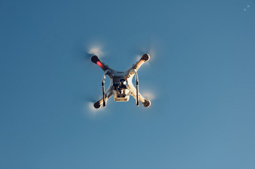 Un drone quadricoptère blanc vole sur un ciel bleu clair, avec ses hélices qui tournent et des lumières rouges allumées sur ses bras.